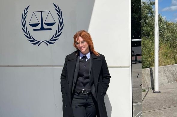 A person in a black suit and trench coat stands in front of a building featuring a picture of the scales of justice.