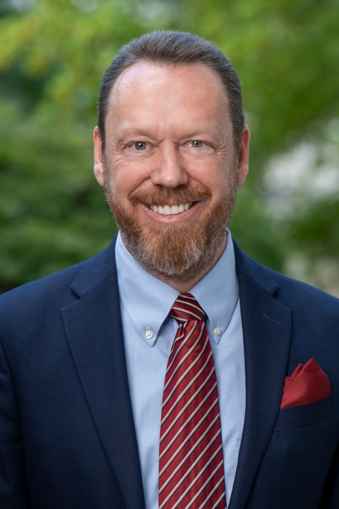 A mean with a beard smiles. He wears a suit and a red tie.