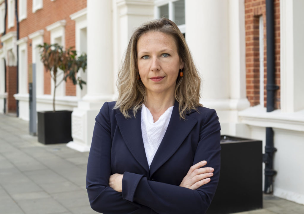 A woman with blonde, shoulder-length hair wears a blue suit jacket and white shirt. She crosses her arms as she stands in front of a red brick building with white columns.