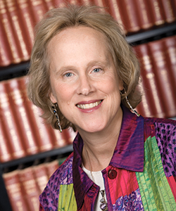 A blonde woman in a colorful shirt smiles in front of a stack of books.
