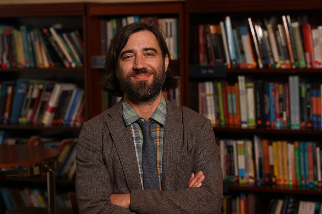 A man wearing a sweater folds his arms in front of shelves full of books