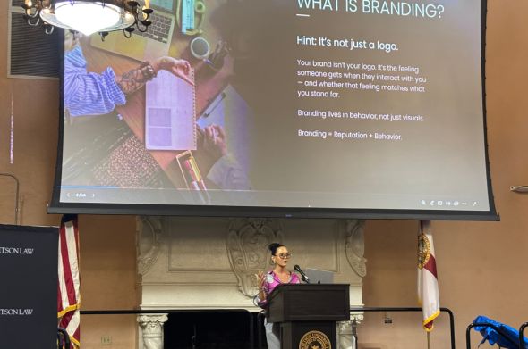 A woman stands at a podium in front of a large screen featuring a slide about branding.