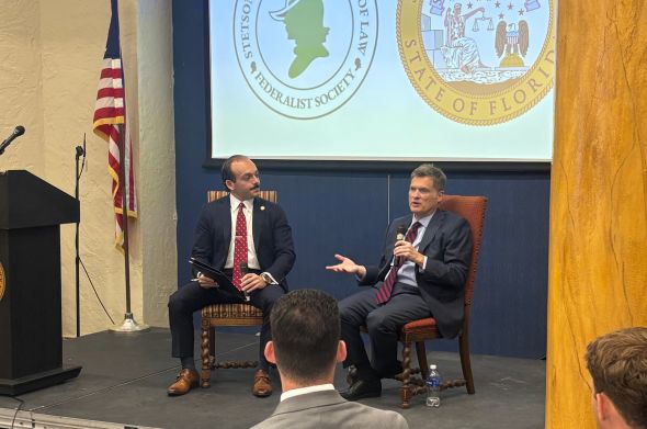 Two men wearing suits and ties sit and talk on a stage in front of a screen displaying the logos for the Stetson Federalist Society and the Florida Supreme Court.