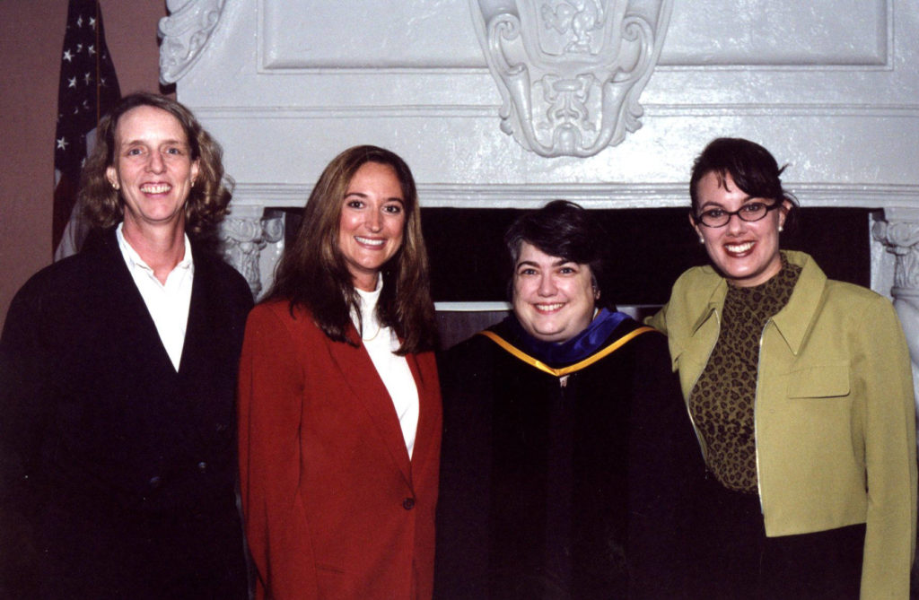 Four women in professional dress smile at the camera in front of a fireplace.