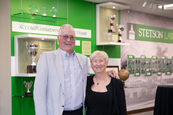 A man wearing a light-colored suit and a woman in a black dress and jacket stand in front of a green wall that is lined with trophies