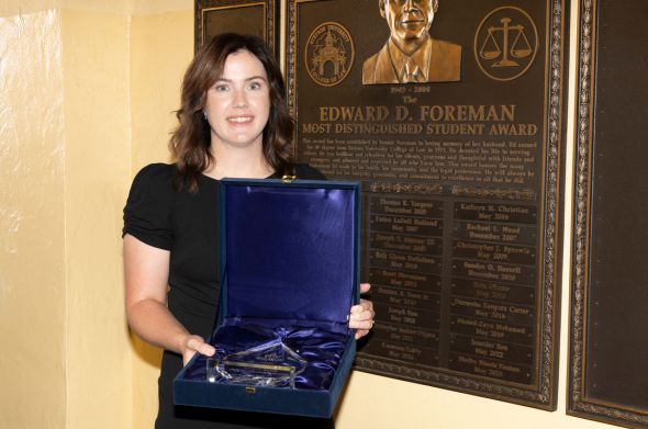 A woman with dark hair and a black dress stands before a plaque highlighting the Edward Foreman Award. She holds the award in a black case lined with purple velvet.