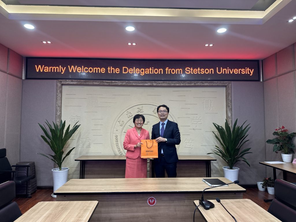 A woman wears a salmon-colored suit and a man wears a dark suit. Together, they hold an orange bag that says "aurora." There are Chinese characters on the wall behind them.