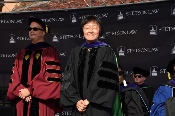 A woman with a dark bob and glasses smiles; she wears full ceremonial regalia. A man in red graduation regalia is in the background.
