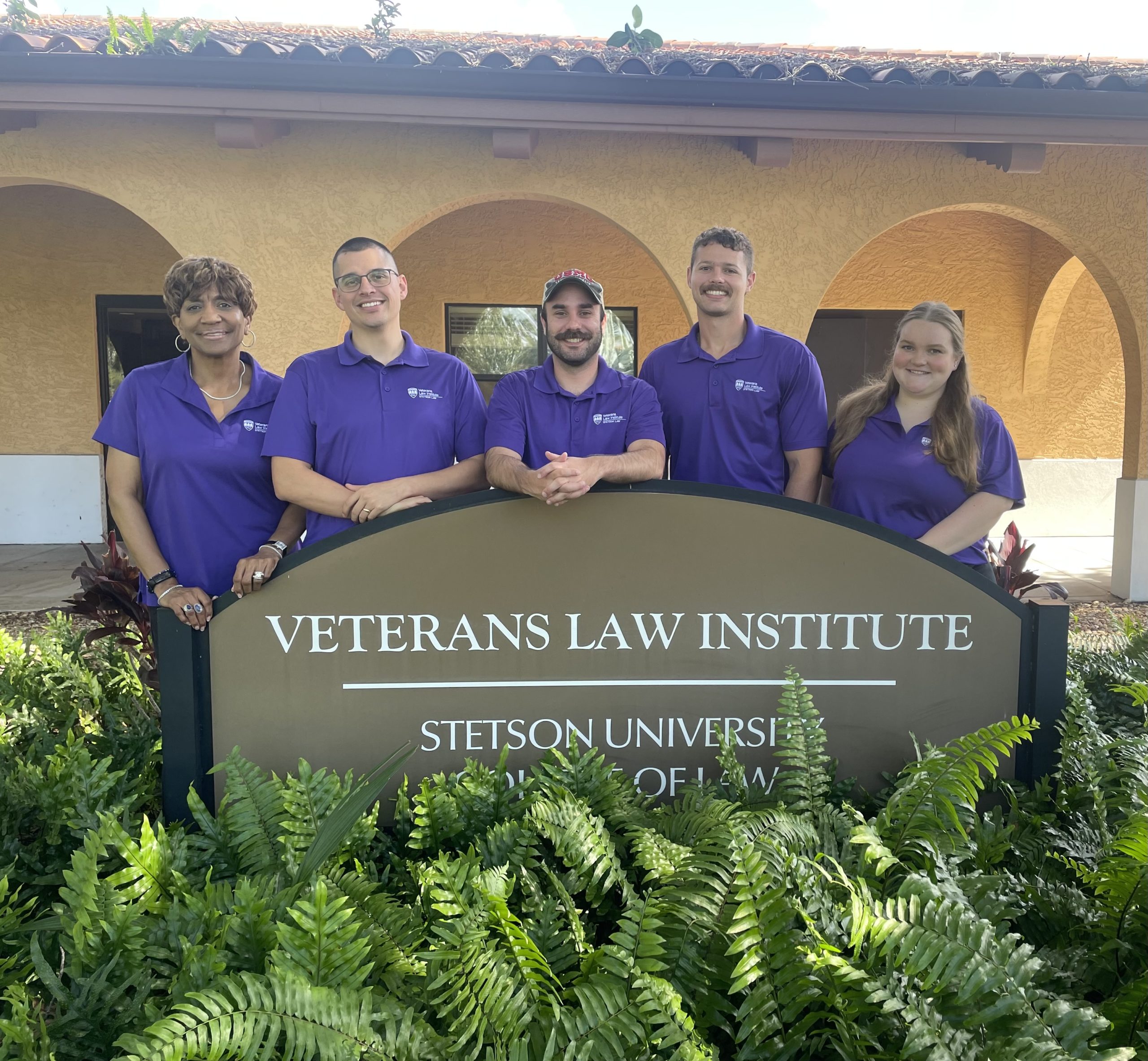 A group of students wearing purple polo shirts stand in front of an outdoor sign that says "VETRANS LAW INSTITUTE"