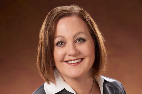 A woman with chin-length brown hair wears a grey suit jacket and white shirt as she smiles for the camera.
