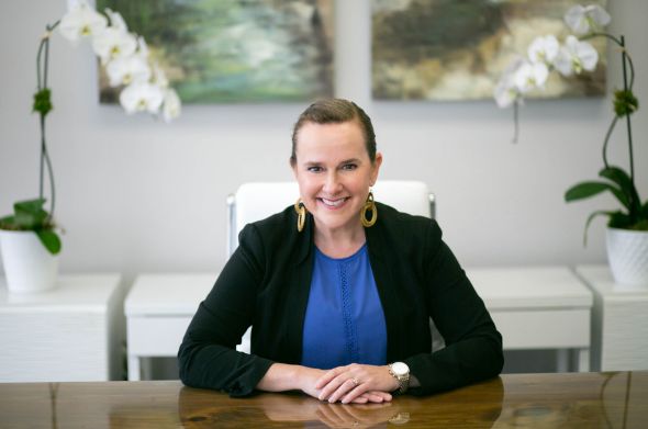 Smiling woman in a blazer sits at a desk in a modern office with orchids and abstract art behind her.