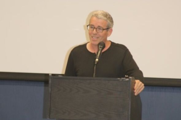 A man wearing a black shirt and glasses speaks at a podium.