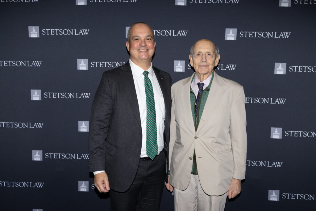 A man in a dark suit and a green tie stands next to a man in a light suit and a dark tie with a green vest in front of a backdrop that says "Stetson Law."