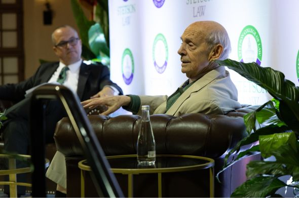 Two men sit on a stage. One is in the foreground, one is in the background.