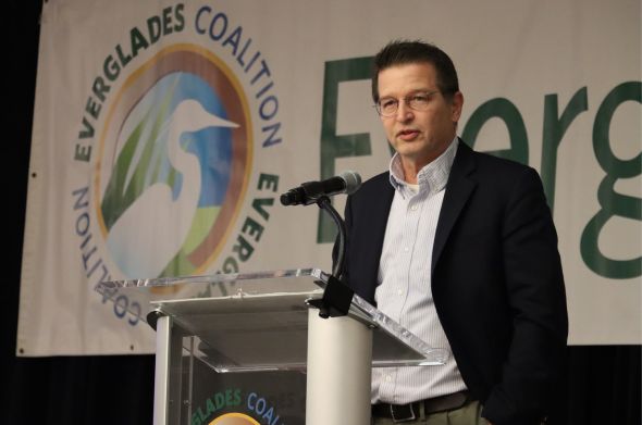 A man with a white shirt and dark suit jacket speaks at a podium in front of a large banned featuring the logo of the Everglades Foundation.