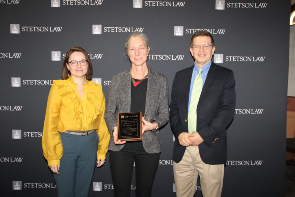 A woman in a gold shirt, a woman in a grey suit jacket holding a plaque, and a man in a black suit jacket stand in front of a backdrop that reads "Stetson Law."