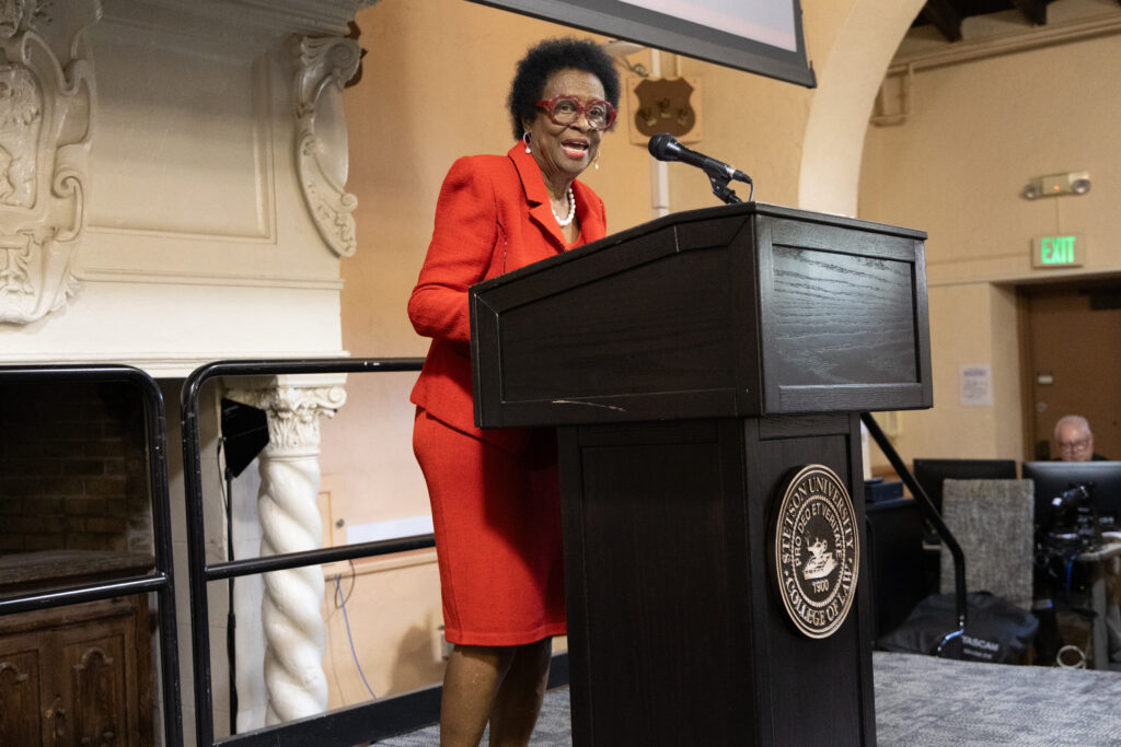 A woman with short hair and a red suit stands at a podium.
