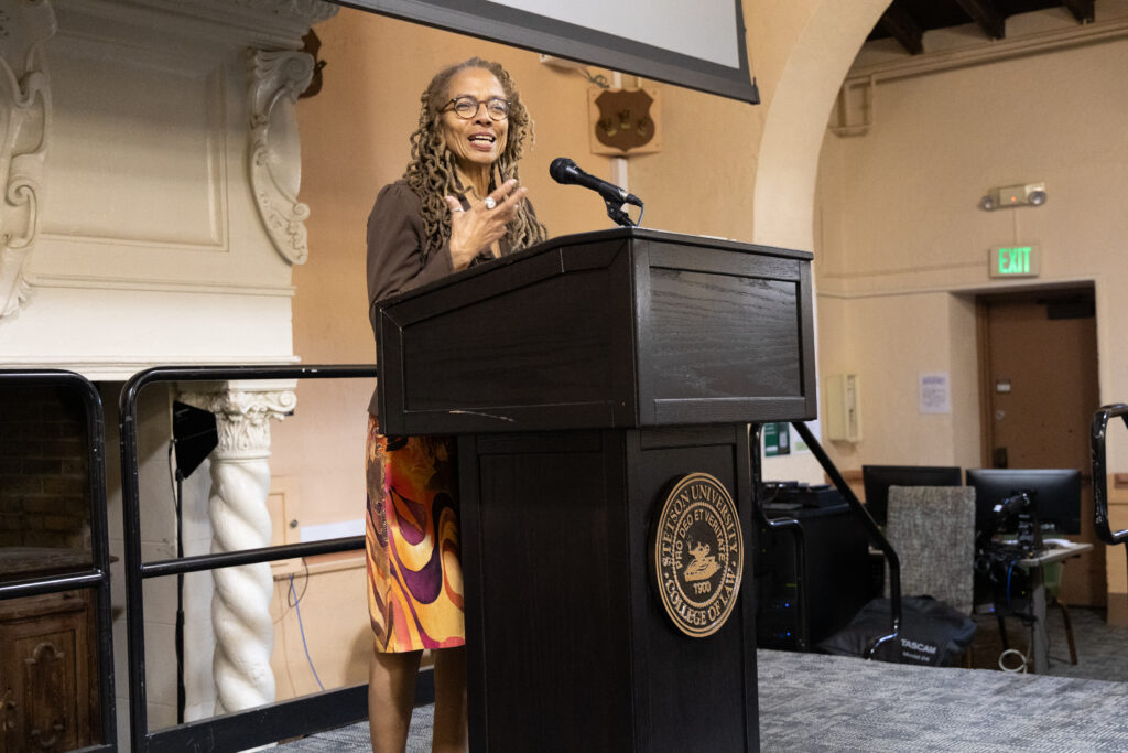 A woman wearing a brown jacket and colorful skirt speaks at a podium.