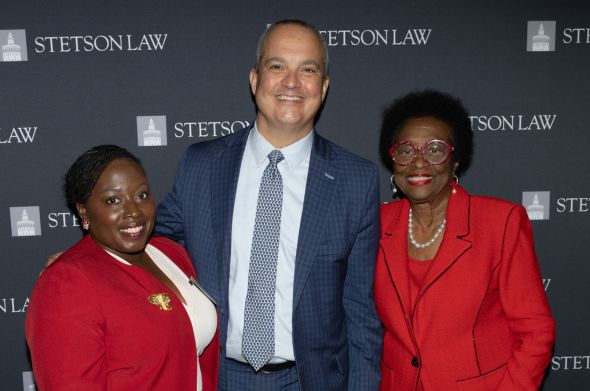 A tall man in a grey suit stands between two women, each wearing red suit jackets. They are in front of a backdrop that reads "Stetson Law."