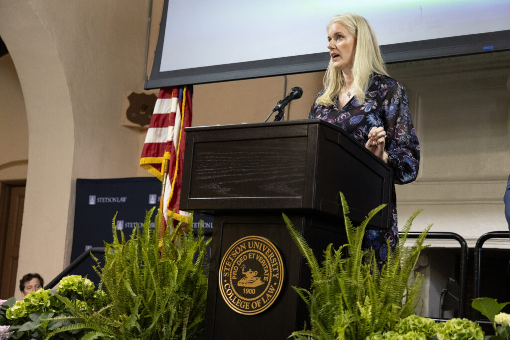 A woman with long blonde hair stands at a podium.