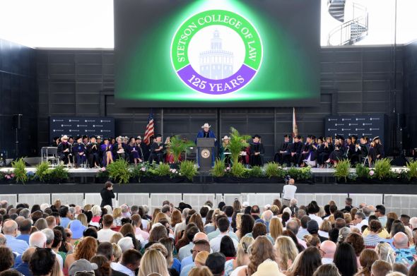 A man in a cowboy hat and graduation robe speaks from a large stage. A big crowd is gathered in audience seating.
