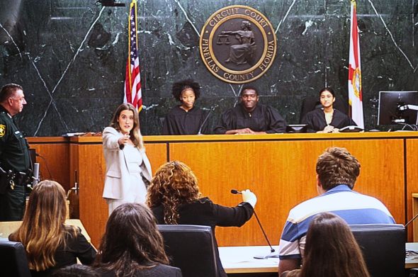 A woman in a white suit stands at the front of a courtroom points toward the camera. Three young students dressed in judges' robes sit along a dais behind her.