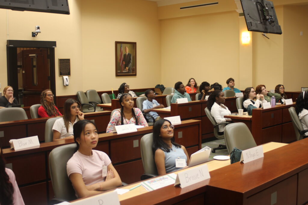 A group of high school students pays attention to a lecture that is taking place in a practice courtroom.