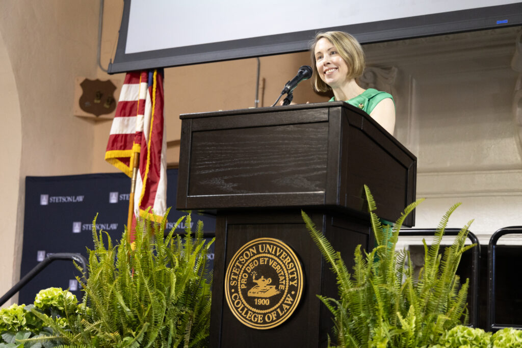 A woman wearing a green dress smiles as she stands behind a podium surrounded by plants on either side.