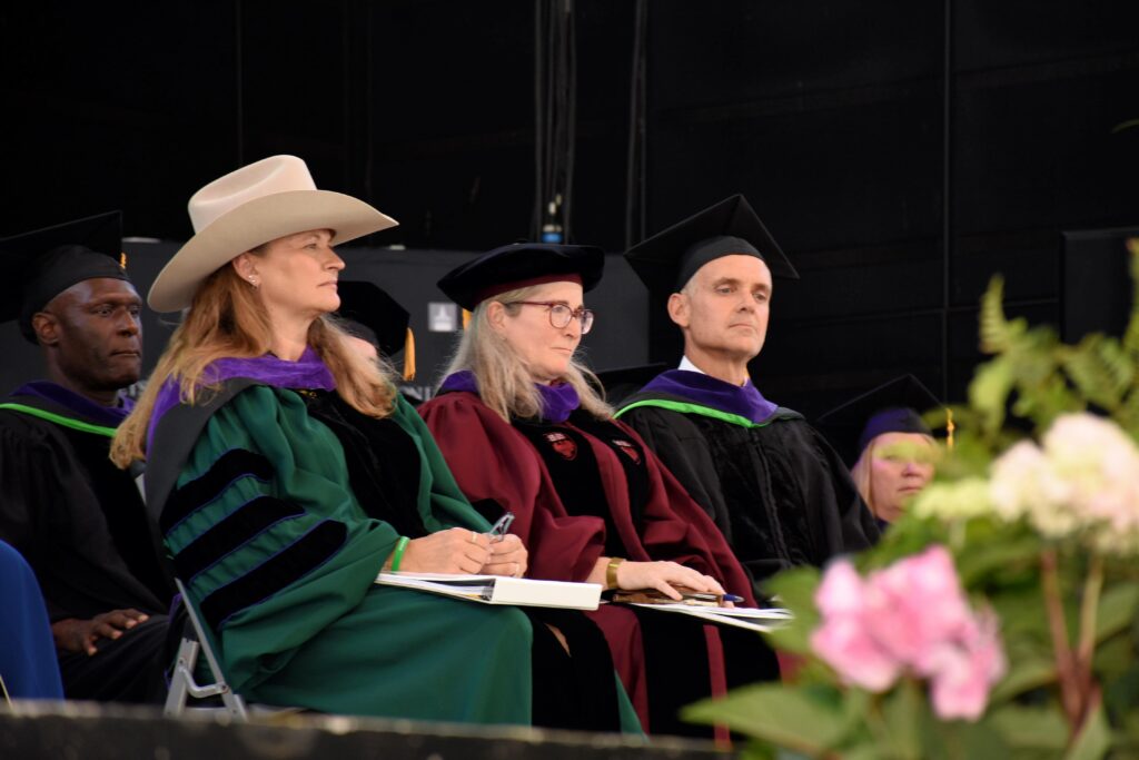 Three people in graduation regalia sit on a stage and observe a commencement ceremony.