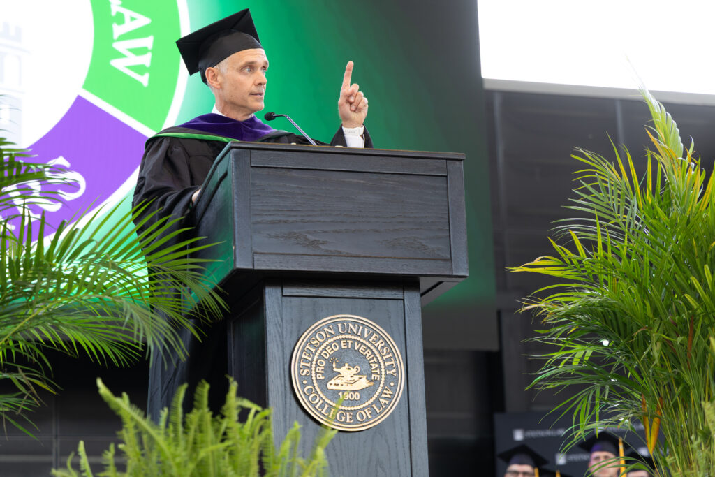 A man in graduation regalia holds his finger up as he speaks at a podium.