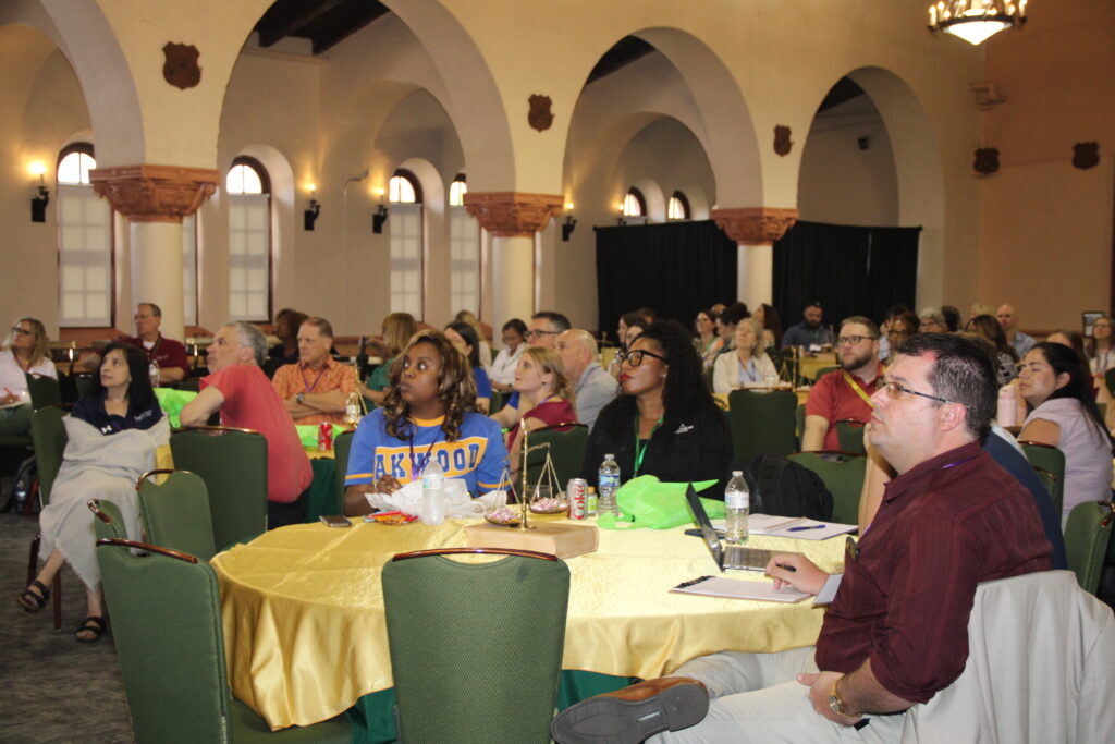 Attendees of a conference of multiple ages and diverse backgrounds are seated at tables in a large event hall.
