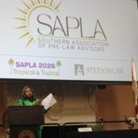 A young woman in a green suit speaks at a podium. The screen behind her reads "S.A.P.L.A."