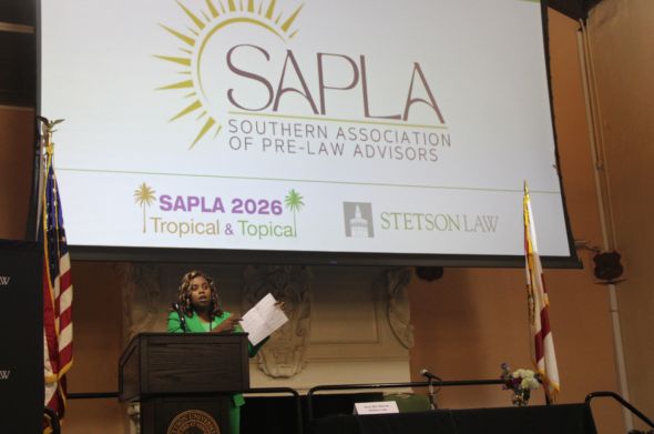 A young woman in a green suit speaks at a podium. The screen behind her reads "S.A.P.L.A."