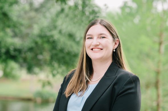 A woman wearing a black suit jacket and light blue shirt smiles. A green, forested area is behind her.