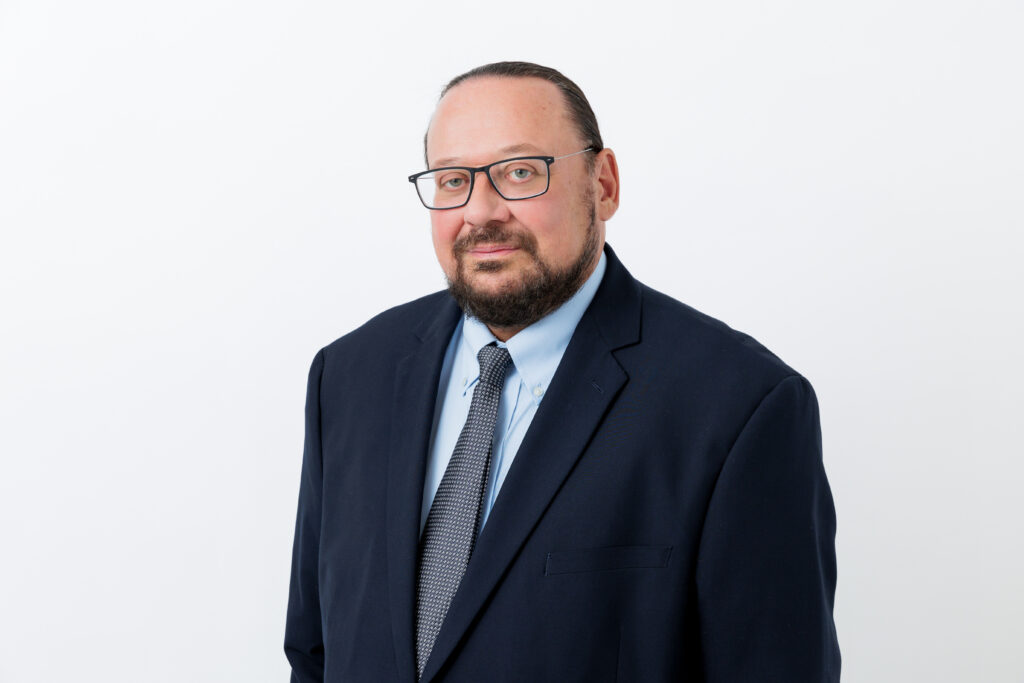 A headshot of a man wearing a suit with a beard and glasses against a white background