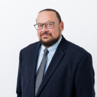 A headshot of a man wearing a suit with a beard and glasses against a white background