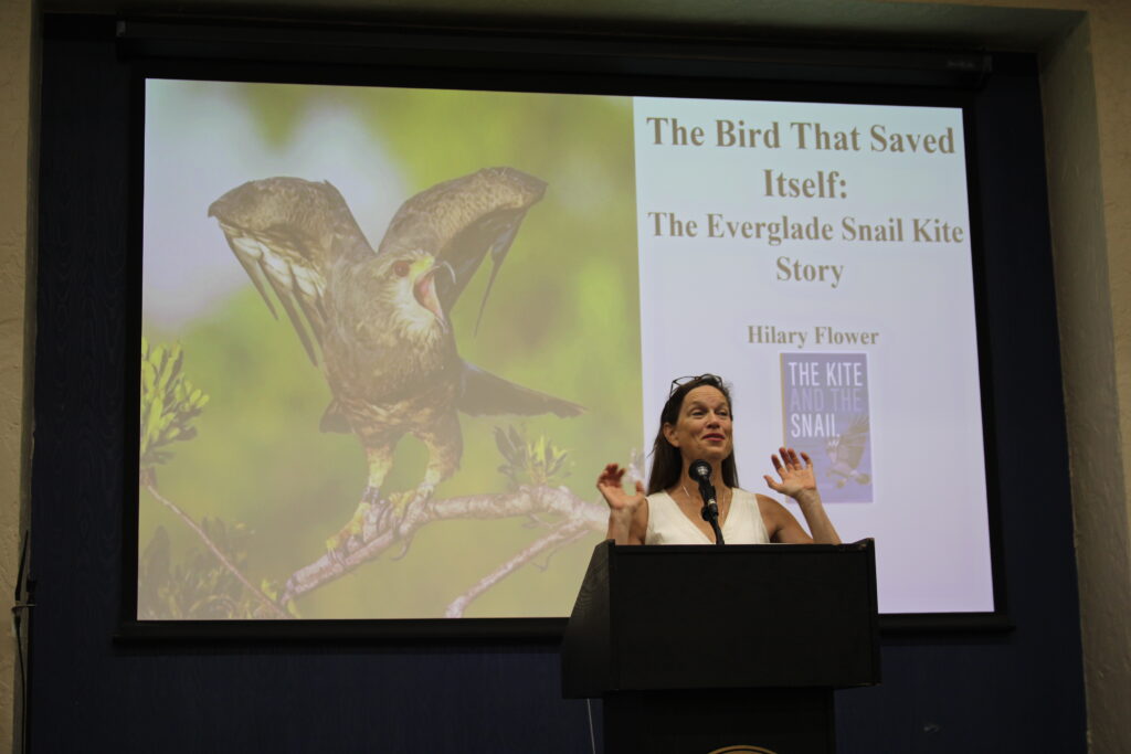 A woman speaks animatedly at a podium. Behind her on a large screen, a projected slide features a bird of prey and the words "The Bird That Saved Itself."