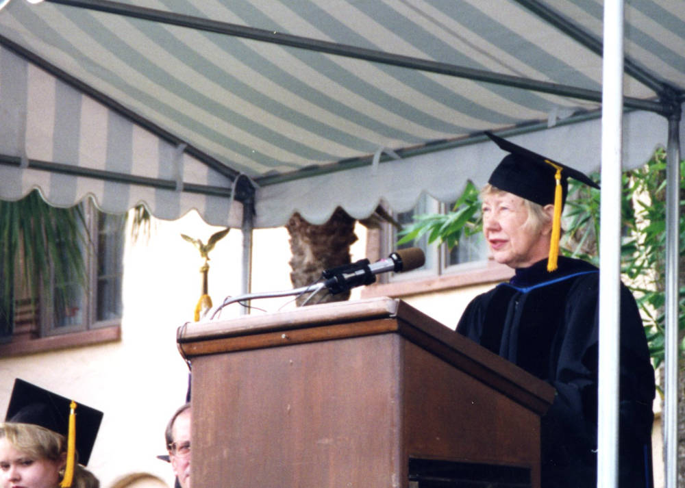 Dean Lizabeth A. Moody wears a cap and gown and speaks to a graduating class.
