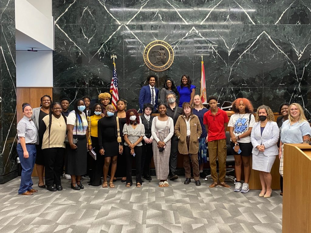 A group of students and mentors take a group shot in a courtroom.