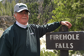 Richard Libby wear Stetson cap and pullover, standing next to Firehole Falls sign.
