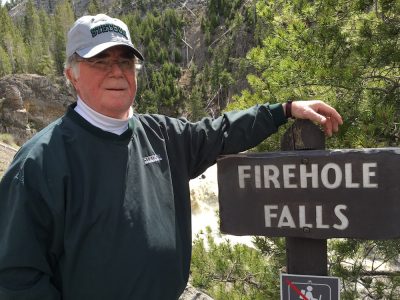 Richard Libby wear Stetson cap and pullover, standing next to Firehole Falls sign.
