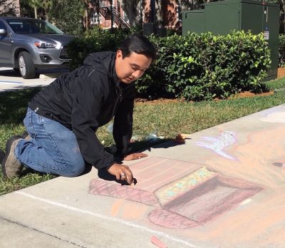 Justin Corriss kneels down on the concrete walkway with colored chalk, drawing a treasure chest.