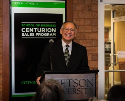 Neal Mero stands smiling and talking at a podium at ribbon cutting.