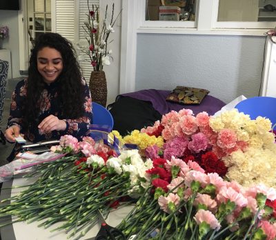 Leah Bayat sits at a table with a pile of colorful carnations, working with construction paper and a hole punch.