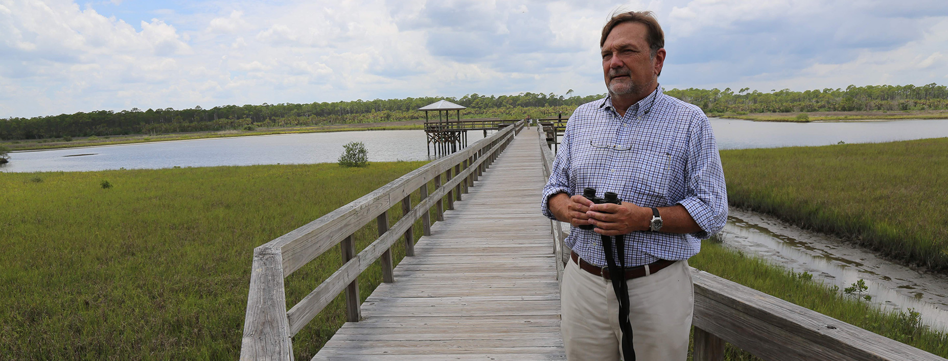Clay Henderson stands in Spruce Creek Park with pretty river and wooden walkway behind him.