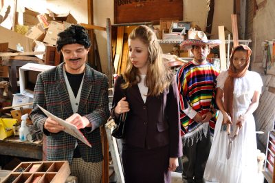 An actor with a cheesy fake moustache shows a woman in a business suit something in a book; while behind them a man in a Mexican garb and a woman watch