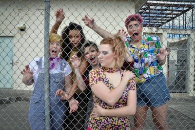 A woman stands with her arms wrapped around herself and her eyes closed as four women make a variety of funny faces behind the fence.
