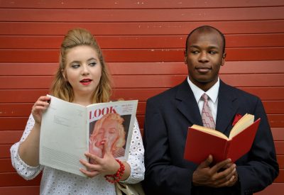 A woman reading a magazine makes eyes at a man standing next to her, against a wall, and he's looking back intrigued.
