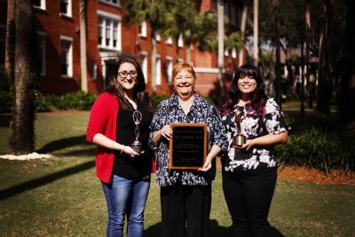 The three stand in front of Elizabeth Hall at Stetson, standing side by side, and holding awards and the plaque.