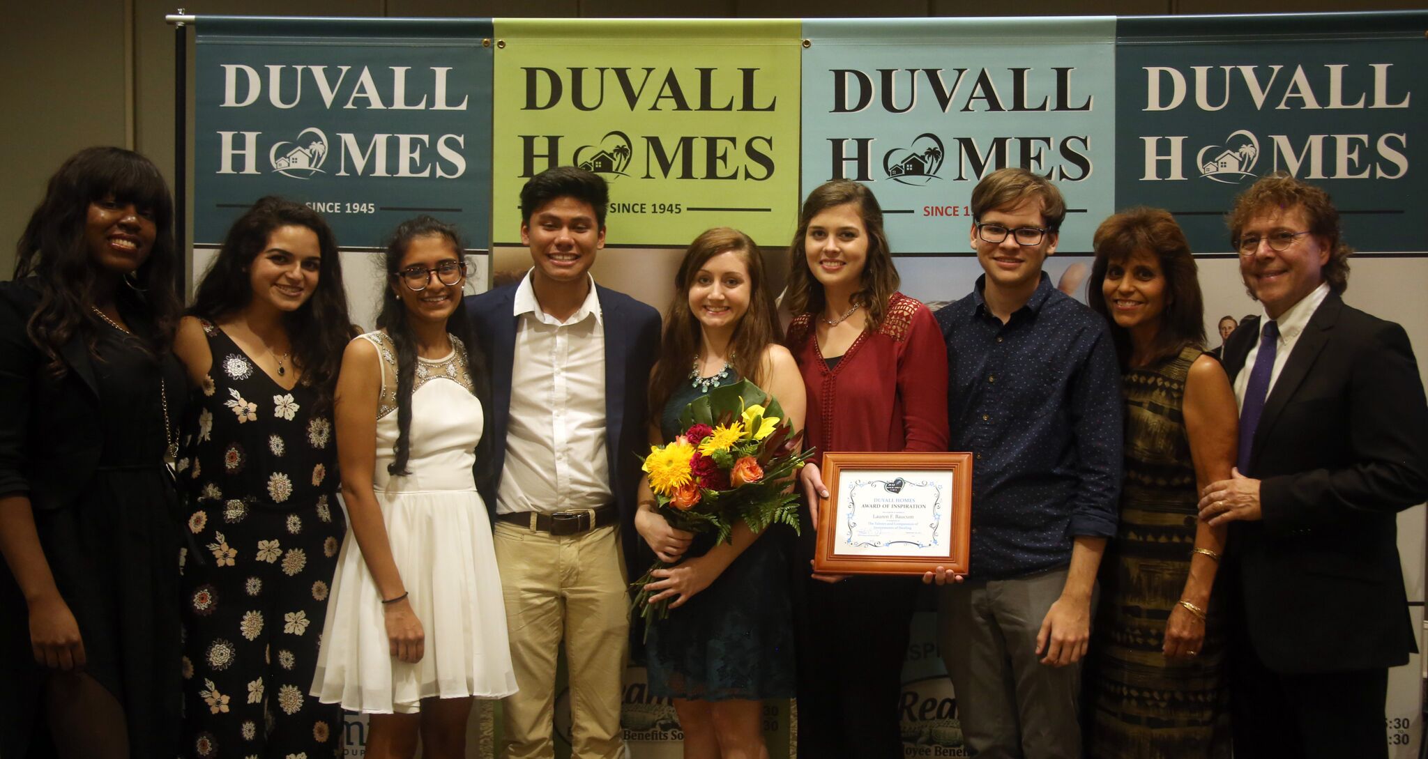 The students, the professor and his wife pose side by side with the framed certificate and a bouquet of flowers, and Duvall Homes printed logos behind them.